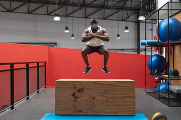 Fit african american man wearing face mask jumping on wooden plyo box while endurance training in th