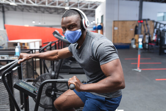 Portrait of fit african american man wearing face mask and headphones exercising on stationary bike 