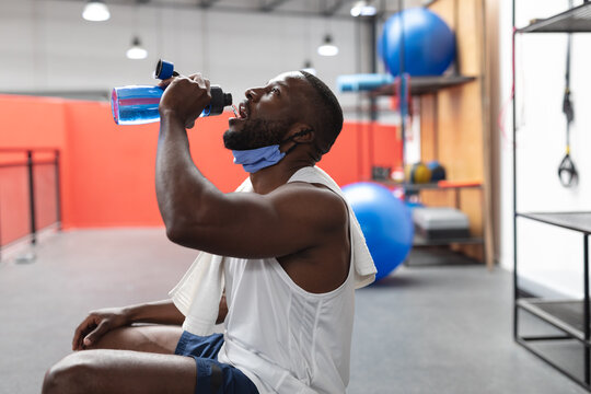 Fit african american man with face mask around his neck drinking water in the gym