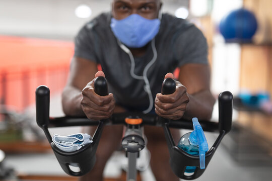 Portrait of fit african american man wearing face mask and earphones exercising on stationary bike i