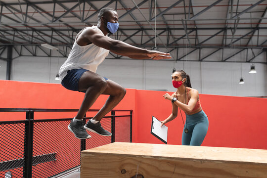 Fit african american man wearing face mask jumping on wooden plyo box in the gym  while  caucasian f