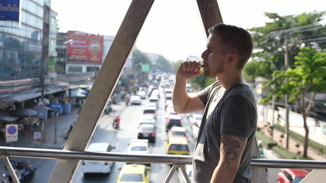Portrait of handsome bearded guy blowing smoke from e-cigarette outdoors. Closeup hipster man smoking electronic cigarette in urban background. slow motion