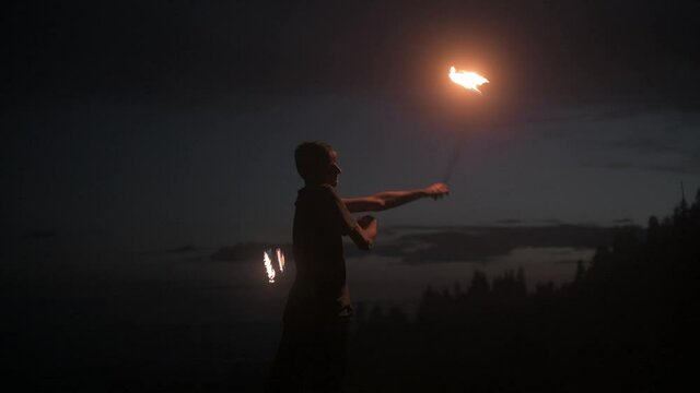 Graceful male fireshow performer making fire dance with two metal fans lit with flames on night mountains meadow