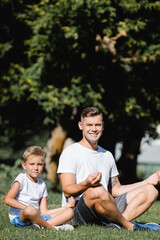 Fototapeta premium Father and son in sportswear looking at camera while relaxing, sitting in lotus pose on grass on blurred background