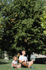 Father and son with closed eyes relaxing while sitting in lotus pose on grass with big tree on background