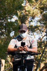Naklejka premium Young man wearing a medical mask using his cell phone sitting on his bike in the forest.