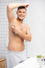 Obraz premium Cheerful shirtless man looking at camera while spraying antiperspirant in bathroom