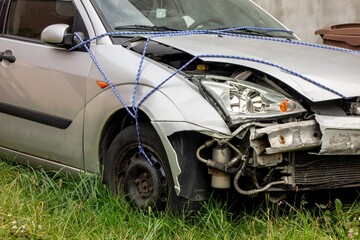 A detail of a frontal part of a silver car damaged in a serious traffic accident with a broken bumper, hood, headlamp which is fixed by rope