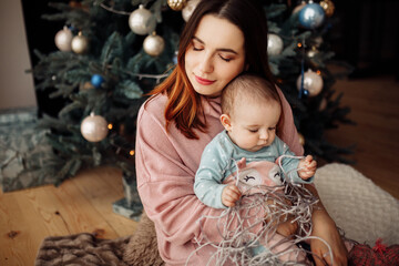 Pretty woman with lovely toddler sit near Christmas tree, tender mother hold cute daughter in arms,...