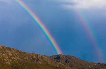 Panorama view of full double rainbow with blue sky and mountain range in background. Near Saint Florent, Corsica