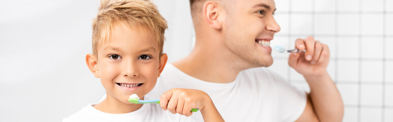 Smiling son with toothbrush looking at camera and standing near father looking away, while brushing teeth in bathroom, banner