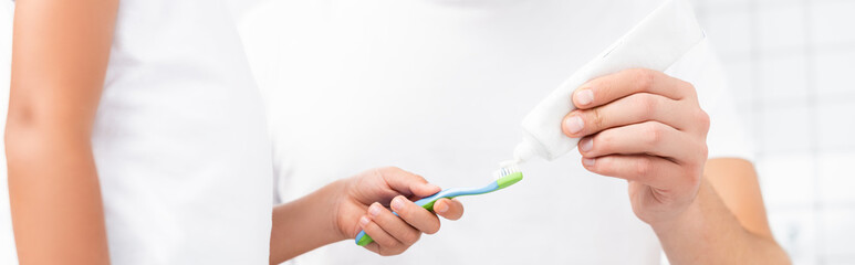 Cropped view of child holding toothbrush while standing near man squeezing toothpaste, banner