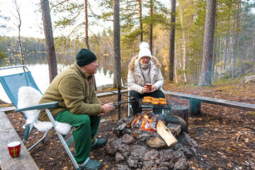 A man and a woman are grilling meat on a fire. Autumn evening outdoors in the forest by the lake. Solitude concept
