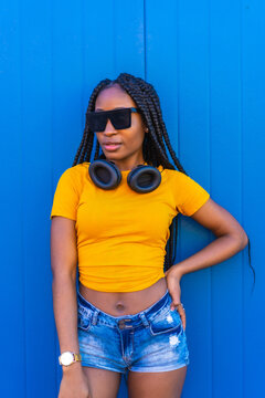 Lifestyle, Black Girl With Long Braids, In A Yellow Shirt And Sunglasses On A Blue Background. Dj With Headphones Smiling Promo Poster Background, Techno, House Music