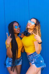 Lifestyle, black girl and blonde Caucasian girl in yellow dresses on a blue background. Very happy and smiling with music headphones and sunglasses