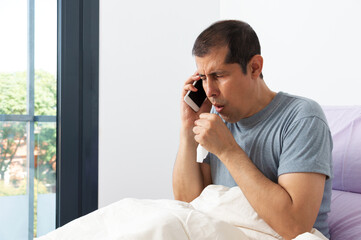 Photo of a sick and coughing businessman sitting on the bed and calling the doctor in the bedroom