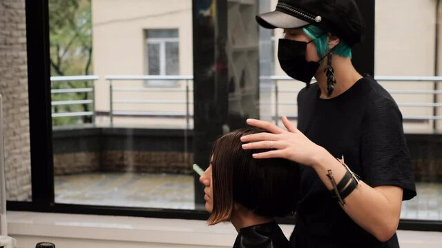 A professional girl hairdresser in face mask makes a client trendy stylish short hair cut. The girl is sitting in stylish beauty salon. Social distance. Protective measures.     