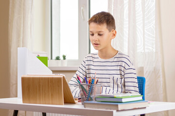 Kid with tablet computer sitting at table with books and having video call, virtual online leasson at home