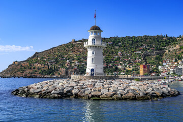 Fototapeta na wymiar Alanya lighthouse on the background of a peninsula with an ancient fortress and modern buildings on the coast (Turkey). Seascape with famous landmarks of the resort - view from the Mediterranean Sea