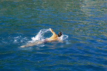 Fototapeta premium A man swims in a bright blue-green sea water. Naked male silhouette waving his hands, splashing water drops. Summer sports background with copy space
