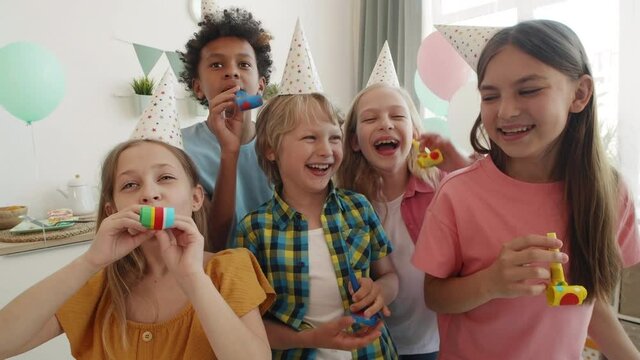 Medium POV of diverse school boys and girls wearing party hats standing indoors. Children smiling, laughing, having fun, using blow ticklers on camera