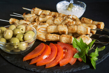 skewer of squid fillet on metal skewers on a dark background with sauce, olives and sliced tomatoes