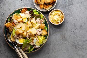 Fresh Caesar salad with lettuce salad, chicken breast, boiled eggs and croutons in ceramic bowl with dressing on the side on gray table. Classic healthy salad, top view, space for text