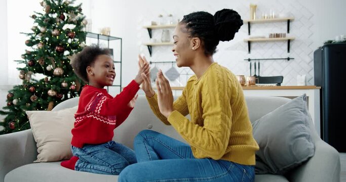 Portrait of happy beautiful African American woman playing with little kid while sitting on sofa in christmassy decorated room Side view of loving mom and child spending time together Holidays concept