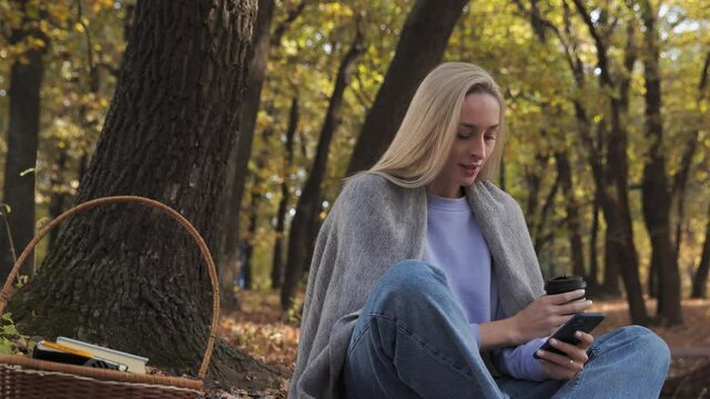 Blonde woman drinks coffee and uses phone in the park at fall on a sunny day