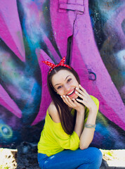 A stylish teenage girl in a yellow jacket and blue jeans poses near a graffiti wall