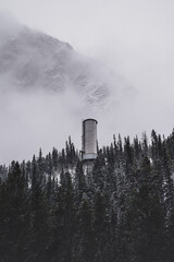 Water Tower Above Forest With Mountain Looming Above It The Clouds 