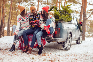 Happy friends in christmas sweaters on a winter walk sit on the trunk of a pickup truck.