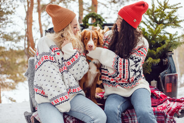 Girls in Christmas sweaters sit with a dog on the trunk of a pickup truck. Car with a Christmas...