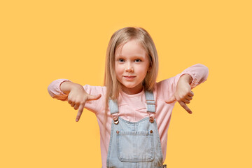 The little blonde girl points her hand down. Yellow isolated background.