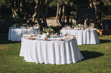 Large round tables with a white tablecloth and empty dishes, a bouquet of flowers stand on green grass in a park in nature. Wedding ceremony, party. Festive decorations, server room.