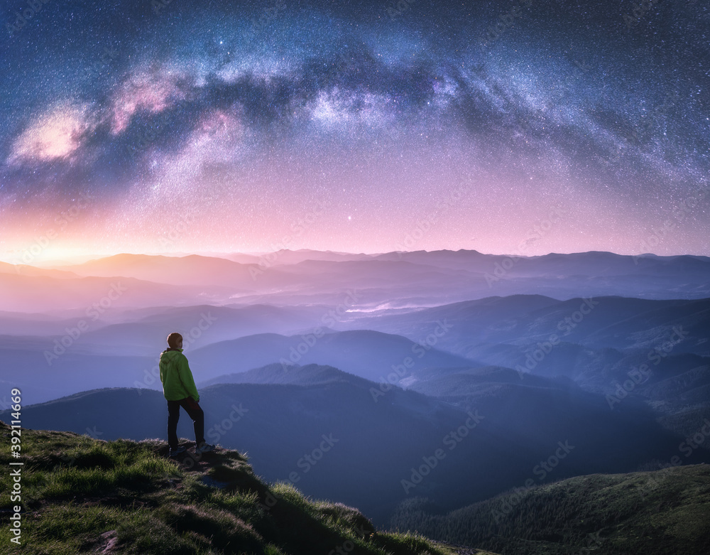 Wall mural man on the mountain peak and arched milky way over mountains in low clouds at night. landscape with 