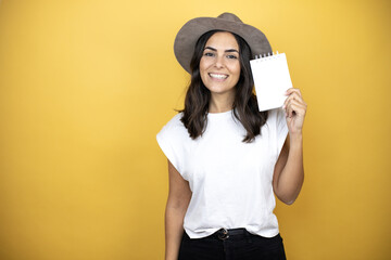 Naklejka premium Beautiful woman wearing casual white t-shirt and a hat standing over yellow background smiling and showing blank notebook in her hand