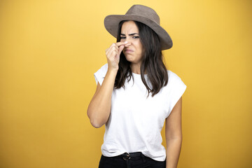 Beautiful woman wearing casual white t-shirt and a hat standing over yellow background smelling something stinky and disgusting, intolerable smell, holding breath with fingers on nose