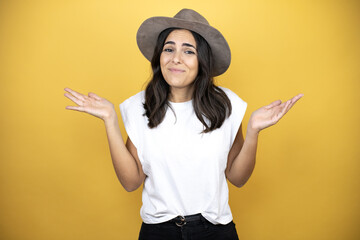 Beautiful woman wearing casual white t-shirt and a hat standing over yellow background clueless and...