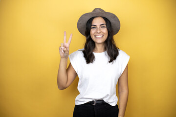 Beautiful woman wearing casual white t-shirt and a hat standing over yellow background showing and pointing up with fingers number two while smiling confident and happy