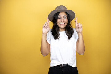 Beautiful woman wearing casual white t-shirt and a hat standing over yellow background gesturing finger crossed smiling with hope and looking side