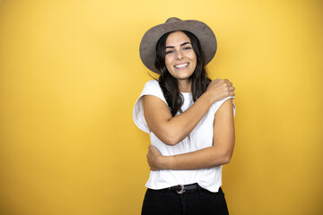 Beautiful woman wearing casual white t-shirt and a hat standing over yellow background hugging...