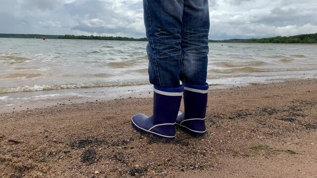 A boy in blue rubber boots standing on a lake shore sand, ye looks on beautiful water landscape with waves