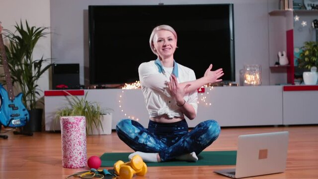 A woman sitting on the floor stretches her back muscles before a sports workout. She twists her body, stretches her arms. Back correction, spine tone. In the background a TV set.