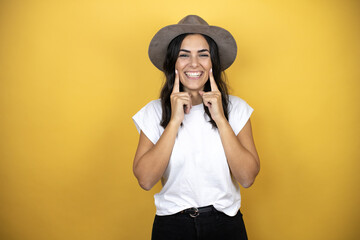 Beautiful woman wearing casual white t-shirt and a hat standing over yellow background smiling confident showing and pointing with fingers teeth and mouth