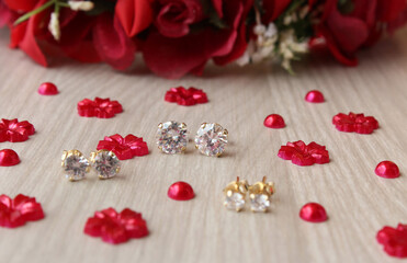 Brincos de ouro com pontos de luz de diamante/zircônia brilhantes sobre fundo de madeira com flores artificiais vermelhas.