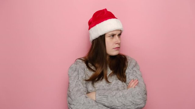 Portrait of sad irritated young woman in Christmas hat looking camera standing with folded crossed hands on chest, wears winter sweater, isolated on pink studio wall. New Year merry holiday concept