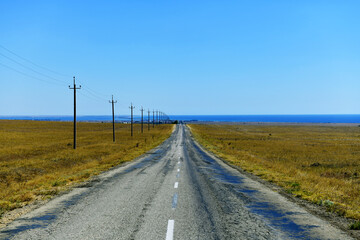 An old asphalt road leading to the sea. In the hot fog, a village in the valley. Dry, sun-scorched grass. Seashore on the horizon. Power poles along the highway.