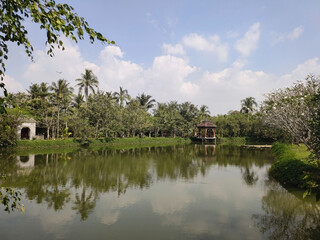 Fototapeta na wymiar Pavilion on the lake. Palm trees, trees, water and sky. Tropical landscape in Ao Dai Museum. Ho Chi Minh City (Saigon). Vietnam. South-East Asia
