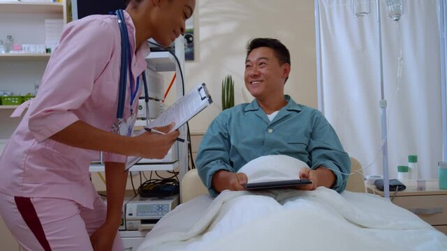 Asian positive man lying in hospital bed welcoming his female nurse communicating about disease recovery. Patient and doctor. Diversity. Healthcare.